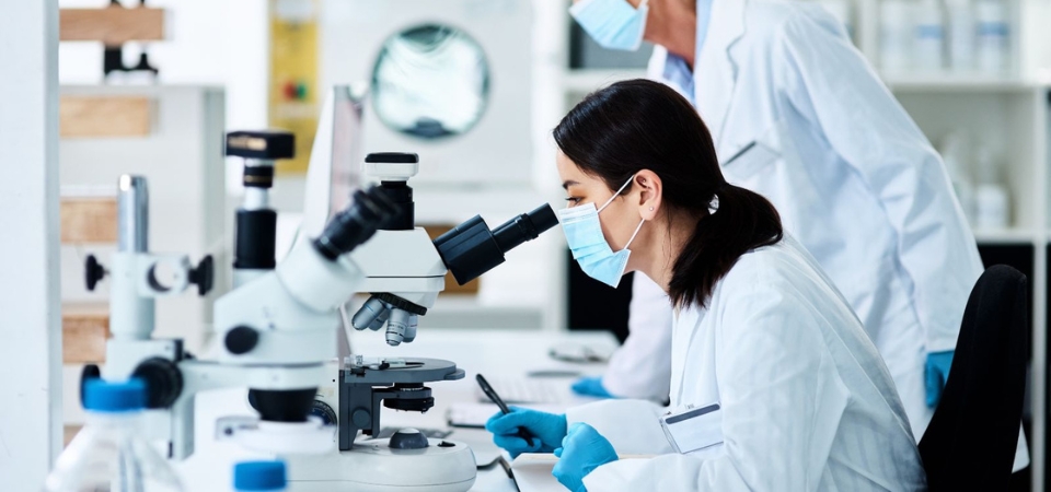Asian woman wearing a hospital face mask, blue gloves, and a white lab coat sits in a lab looking into a microscope. 