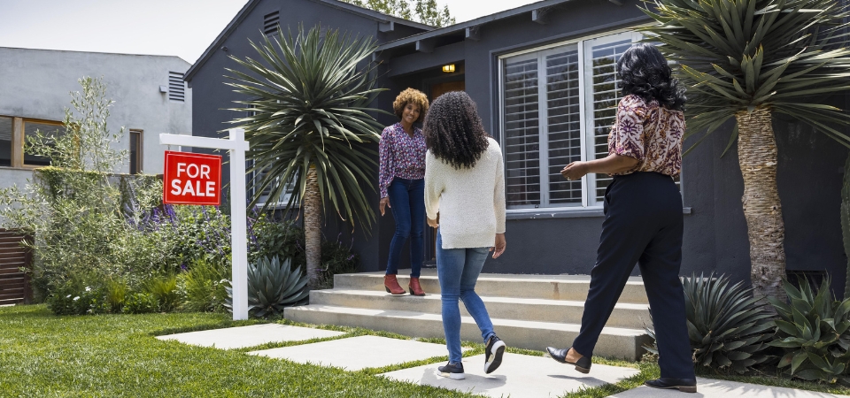 African American women walk on pathway to a house with a "For Sale" sign in the yard. An African American woman, presumably the realtor, stands at the front door smiling.