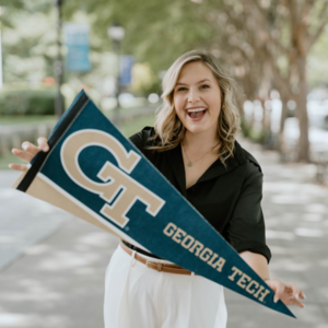 Laura Gregory poses with a Georgia Tech pennant on Georgia Tech's campus. Walkways and trees appear in the background.