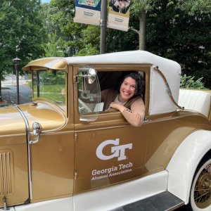 Lauren Kennedy sits in the driver's seat of the Ramblin' Wreck on a brick walkway on Georgia Tech's campus. A lamppost with banners featuring the Transforming Tomorrow campaign appears in the background.