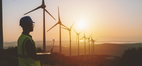 Silhouette of young male engineer holding laptop computer planning and working for the energy industry and standing beside a wind turbines farm power station at sunset time.