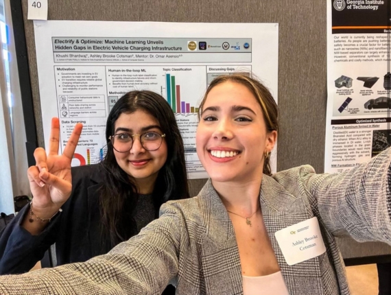 Ashley takes a selfie with a friend in front of a poster presentation at a conference.