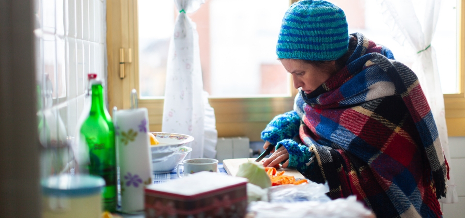 A woman inside an apartment kitchen wearing a beanie, sweater, and gloves while she prepares food.