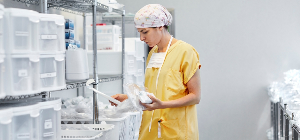 Person in scrubs standing in hospital supply room holding packages of medical supplies.