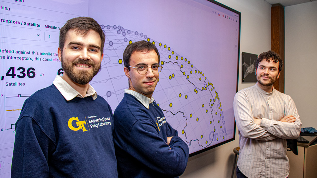 Researchers stand in front of a screen displaying figures and dots that represent space-based interceptor satellites above a map image of Earth.