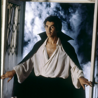 a handsome man in a cloak and open white shirt stands in a doorway, with misty darkness in the background
