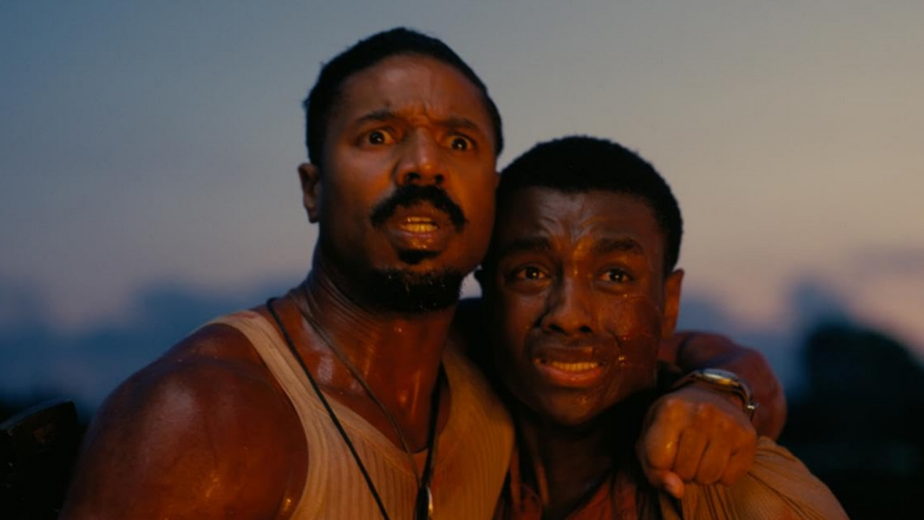 Two men stand together, looking at something behind the camera with expressions of fear. Firelight is reflected on their faces and bodies.