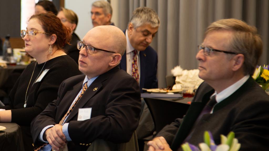 People seated at tables listen to a speaker who is off camera during a symposium