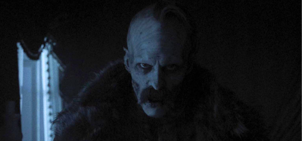 a cadaverous, pale figure wearing a cloak stands in the shadows, with a dim window in the background