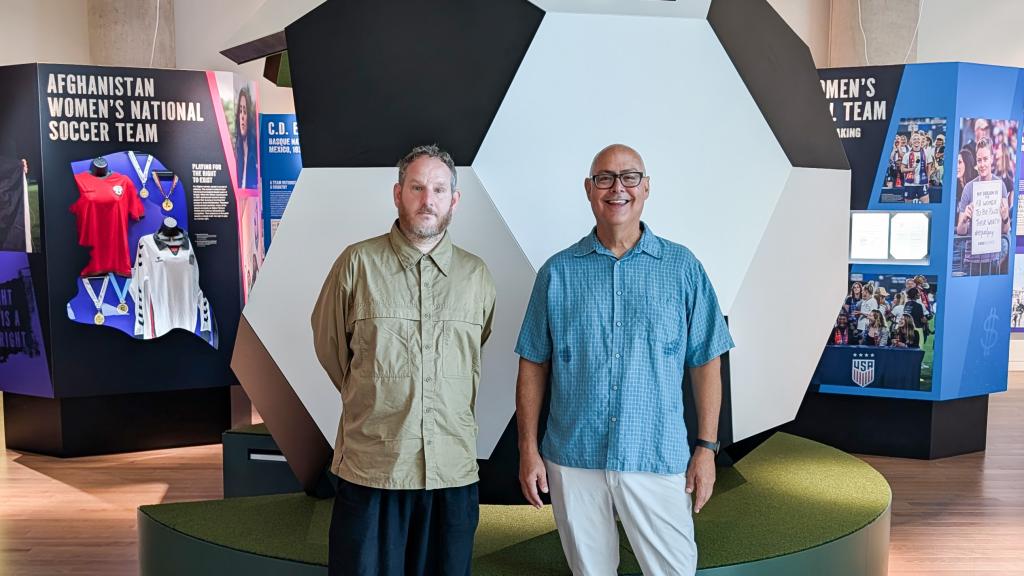 Daniel Fuller and Kirk Bowman stand in front of a large soccer ball and other displays.